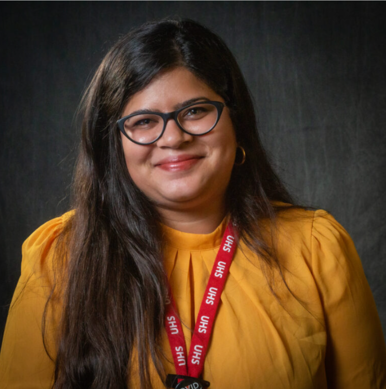 Picture of woman in yellow shirt, glasses, and long brown hair smiling at the camera, in front of a grey background.