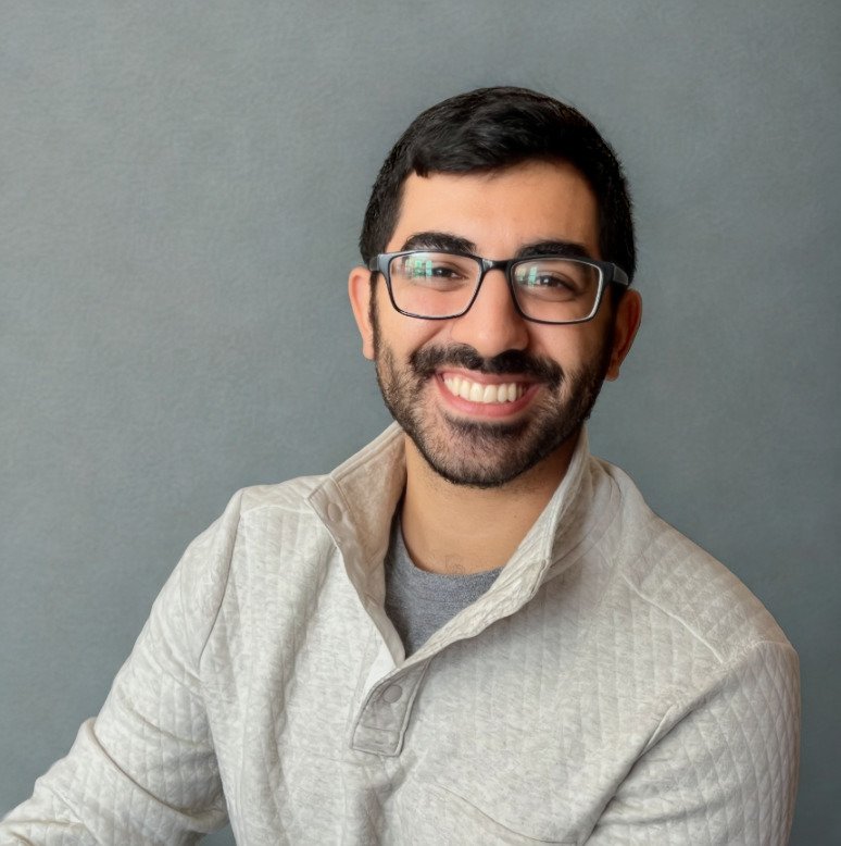 Man with black hair and glasses, smiling at the camera in front of a grey background.