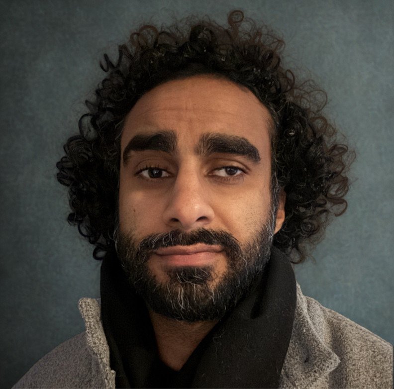 Man with black curly hair and beard looking at the camera, in front of a grey background.