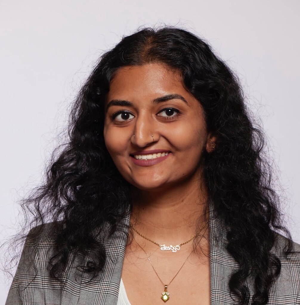Woman wearing a grey suit jacket and with long black wavy hair smiling at the camera.